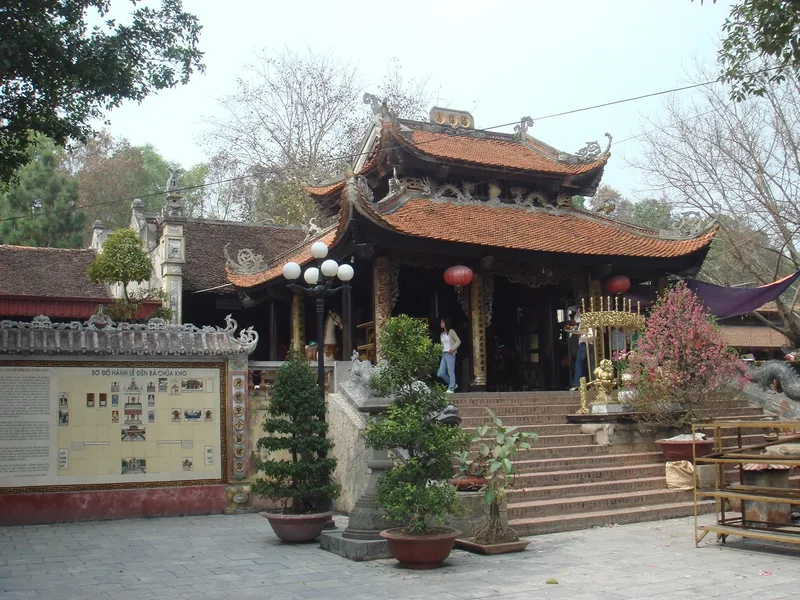 Mái ngói đỏ chạm khắc tinh xảo của đền Bà Chúa Kho ở Bắc Ninh, với chậu cây bonsai và bậc thang đá dẫn vào cửa đền.
